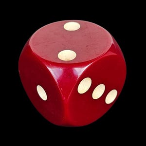 May include: A close-up of a red die with white spots. The die has three visible faces, each showing a different number of spots. The die is a bright red colour and has a glossy finish. The background is black.