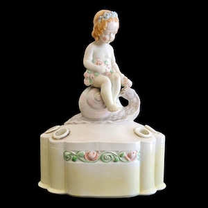 May include: A vintage porcelain figurine featuring a cherubic figure seated on a swan. The figurine is painted in soft pastel colors, with a floral garland and a decorative base with rose accents. The piece has a cream-colored finish.