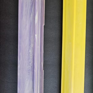 May include: Two long, rectangular strips of material. The left strip features a marbled pattern of light purple and white. The right strip is a solid, bright yellow color. Both items have a raised, central ridge running vertically along their length.