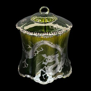 Op de afbeelding: Een groene glazen pot met een deksel, met een zilveren drakenontwerp. De pot heeft een uitlopende vorm en een zilveren rand. Het deksel heeft een kleine lusgreep.