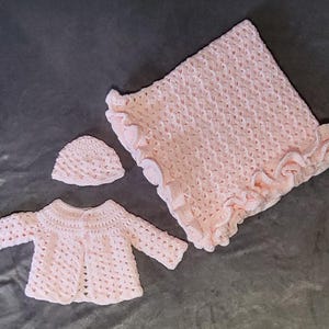 Puede incluir: Un conjunto de bebé de ganchillo rosa claro, que incluye una manta con borde con volantes, un gorro y una rebeca con botones. El conjunto está hecho de hilo suave y se muestra sobre una superficie gris.