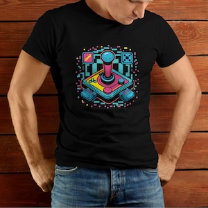 Puede incluir: Una camiseta negra con un gráfico de joystick de estilo retro. El joystick es rosa, azul y amarillo con una base negra. El gráfico está rodeado por un borde pixelado.