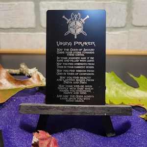 May include: A black metal card with the words "Viking Prayer" engraved in white. The card features a shield and crossed axes design at the top. The prayer is displayed in a stylized font, and the card is propped on a small wooden easel.