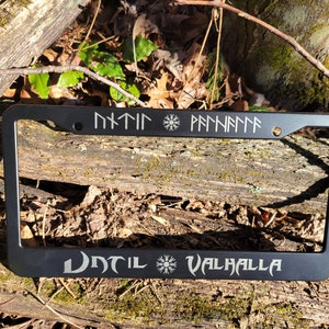 May include: Black metal license plate frame with the text "Until Valhalla" and a snowflake symbol. The top of the frame has runes in the text "Odin" and "Freyja".