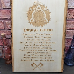 Puede incluir: Un letrero de madera con un código vikingo grabado en él. El letrero presenta una brújula, un triskel y dos hachas cruzadas. El texto dice: "Código Vikingo Protege a tu familia. Honra a los mayores. Enseña a los jóvenes. Sé leal a tu amigo. Di tu opinión. Mantente firme. Toma el mando cuando otros muestren debilidad. Juega cuando puedas. Trabaja cuando debas. Deja siempre tu huella."
