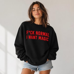 May include: Black crewneck sweatshirt with the red text "F*CK NORMAL I WANT MAGIC". The sweatshirt has long sleeves and a relaxed fit. The model is wearing denim shorts.