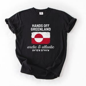 May include: Black t-shirt with the text "HANDS OFF GREENLAND" above a Greenland flag design. Below the flag, the text reads "arctic & atlantic" and coordinates. The shirt is made of a soft material.