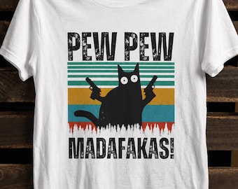 May include: A white short-sleeved t-shirt featuring a graphic of a black cat holding two handguns. Above the cat is the text PEW PEW and below is the text MADAFAKAS! The design includes a retro-style background with horizontal stripes in green, yellow, and blue colors.