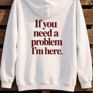 Puede incluir: Una sudadera con capucha blanca que presenta el texto rojo oscuro If you need a problem I'm here impreso en la parte posterior. La prenda incluye capucha, mangas largas con puños acanalados y dobladillo acanalado, colgada de dos pinzas.