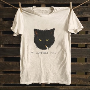 Puede incluir: Camiseta blanca con un gato negro con ojos amarillos y un cigarrillo en la boca. El texto "THE VAMPIRE'S WIFE" está impreso debajo. La camiseta está colgada en un tendedero contra un fondo de madera.