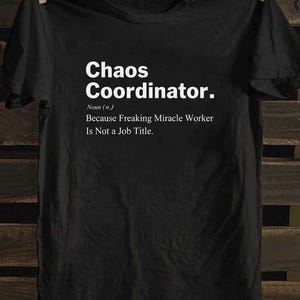 May include: Black t-shirt with the text "Chaos Coordinator." in white, along with the definition: "Noun (n.) Because Freaking Miracle Worker Is Not a Job Title." The shirt is hanging on a wooden background.