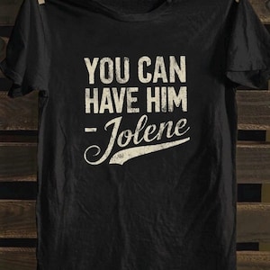 Op de afbeelding: Zwart t-shirt met de tekst "YOU CAN HAVE HIM - Jolene" in verweerde witte letters. Het shirt hangt aan een waslijn tegen een houten achtergrond.