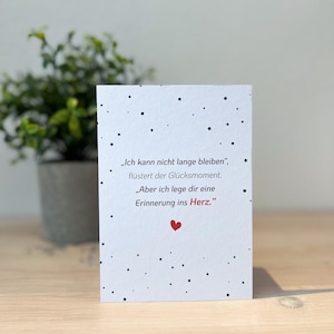 Puede incluir: Una tarjeta blanca con motas negras y texto en alemán. El texto incluye las frases "Ich kann nicht lange bleiben" y "Erinnerung ins Herz". Un pequeño corazón rojo está en la parte inferior. Una planta en maceta está en el fondo.