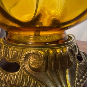 May include: Close-up of a vintage amber glass lamp with a decorative brass base. The base features ornate scrollwork and a detailed design. The lamp's glass shade has a rounded shape, and the brass base adds a touch of elegance.