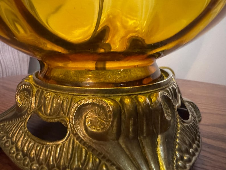 May include: Close-up of a vintage amber glass lamp with a decorative brass base. The base features ornate scrollwork and a detailed design. The lamp's glass shade has a rounded shape, and the brass base adds a touch of elegance.