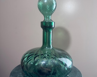 May include: A green glass decanter with a round body and a spherical stopper. The decanter has a textured, cut-glass design. It is displayed on a small, round, green marble-topped table. The decanter is an elegant piece for serving beverages.
