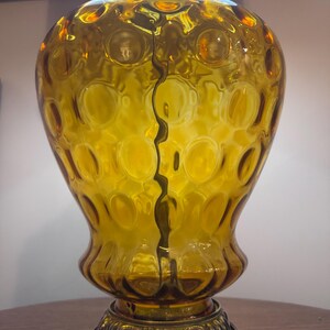 May include: Amber-colored glass table lamp with a textured, bubble-like pattern. The lamp has a decorative metal base and a crack running down the center. The lamp is turned on, and the light is visible.
