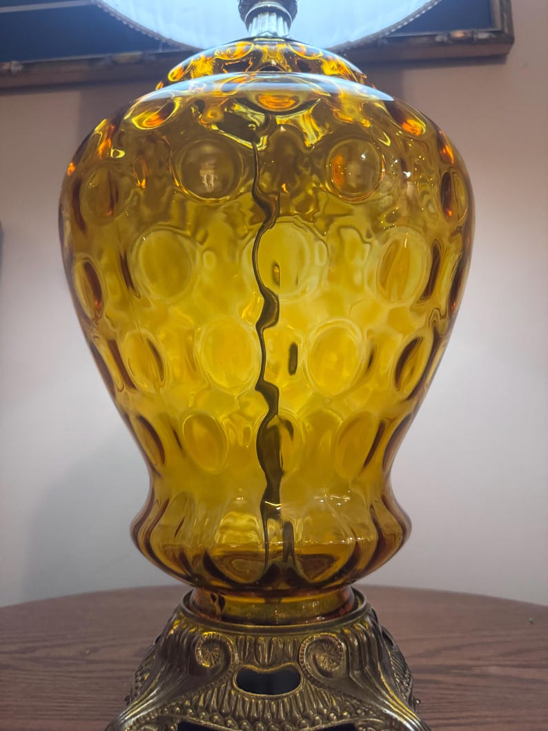 May include: Amber-colored glass table lamp with a textured, bubble-like pattern. The lamp has a decorative metal base and a crack running down the center. The lamp is turned on, and the light is visible.