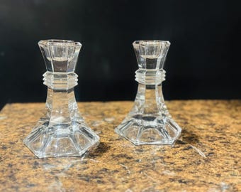 May include: Two clear glass candle holders with a faceted design. The candle holders have a hexagonal base and a flared top to hold candles. The glass is transparent, reflecting light and creating a sparkling effect. The candle holders are on a brown surface.