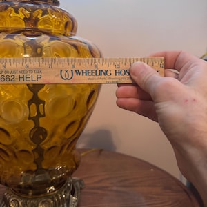 May include: Amber-colored glass table lamp with a decorative base. The lamp has a textured surface with a bubble pattern. The lamp is being measured with a ruler. The text on the ruler says "WHEELING HOS".