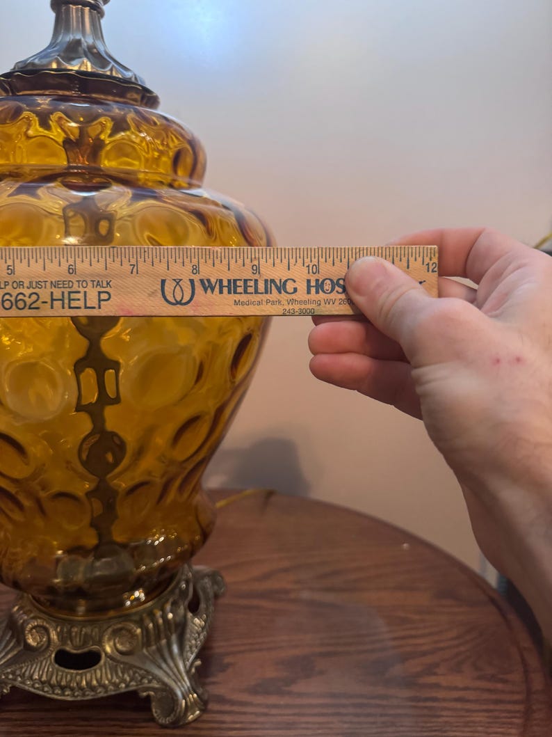 May include: Amber-colored glass table lamp with a decorative base. The lamp has a textured surface with a bubble pattern. The lamp is being measured with a ruler. The text on the ruler says "WHEELING HOS".