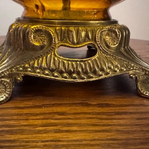 May include: A metallic lamp base featuring an ornate, embossed design with scrollwork and circular motifs. The base supports a translucent amber-colored glass element, showcasing a detailed, textured finish with a warm, golden-toned metal surface.