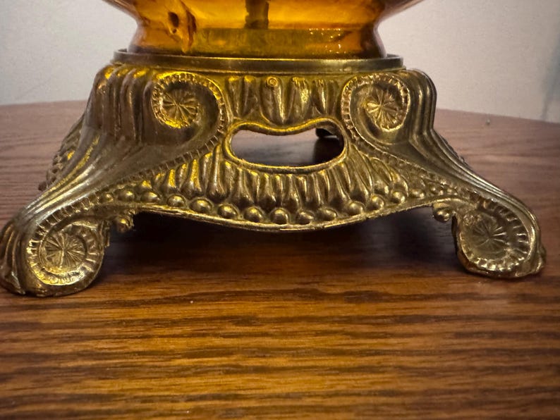 May include: A metallic lamp base featuring an ornate, embossed design with scrollwork and circular motifs. The base supports a translucent amber-colored glass element, showcasing a detailed, textured finish with a warm, golden-toned metal surface.