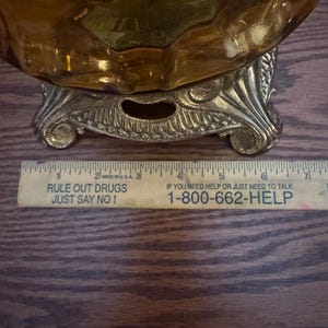May include: An amber-colored glass bowl with a textured surface sits on a decorative gold-toned metal base. A yellow ruler is positioned in front, showing the base width is approximately 4 inches across the bottom edge.