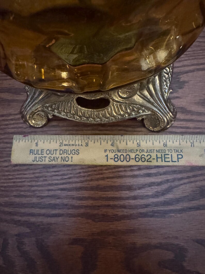 May include: An amber-colored glass bowl with a textured surface sits on a decorative gold-toned metal base. A yellow ruler is positioned in front, showing the base width is approximately 4 inches across the bottom edge.
