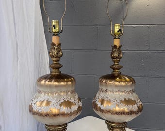 May include: Pair of vintage table lamps with ornate brass bases and accents. The lamps feature a gold and white glass shade with a lace-like design. Each lamp sits on a square marble base. The lamps are wired and ready for use.