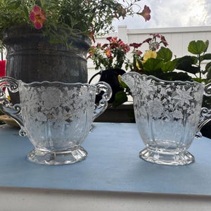 May include: A clear glass sugar bowl and creamer set. Both pieces feature a floral etched design and ornate handles. The sugar bowl has two handles, while the creamer has one. The set is displayed on a light blue surface.