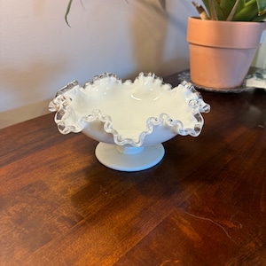 May include: A white glass bowl with a silver rim and a ruffled edge. The bowl is on a pedestal and has a delicate, feminine design.