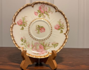 May include: A white porcelain saucer with a gold rim and pink rose floral pattern. The saucer has a scalloped edge and is on a wooden stand.