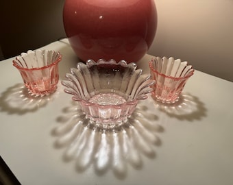 May include: Three pink glass bowls with a scalloped edge. The bowls are arranged on a white surface.