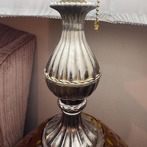May include: A vintage table lamp with a silver-colored, ribbed metal base and a decorative amber-colored glass bottom. The lamp features a white lampshade and a pull chain. The lamp's base has a decorative rope-like detail.