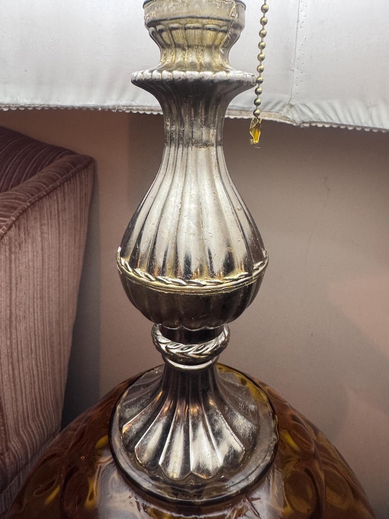 May include: A vintage table lamp with a silver-colored, ribbed metal base and a decorative amber-colored glass bottom. The lamp features a white lampshade and a pull chain. The lamp's base has a decorative rope-like detail.