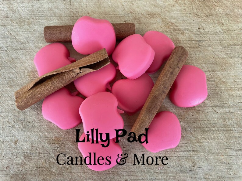 May include: A close-up of pink apple-shaped wax melts with cinnamon sticks on a wooden surface. The text "Lilly Pad Candles & More" is visible in the image.