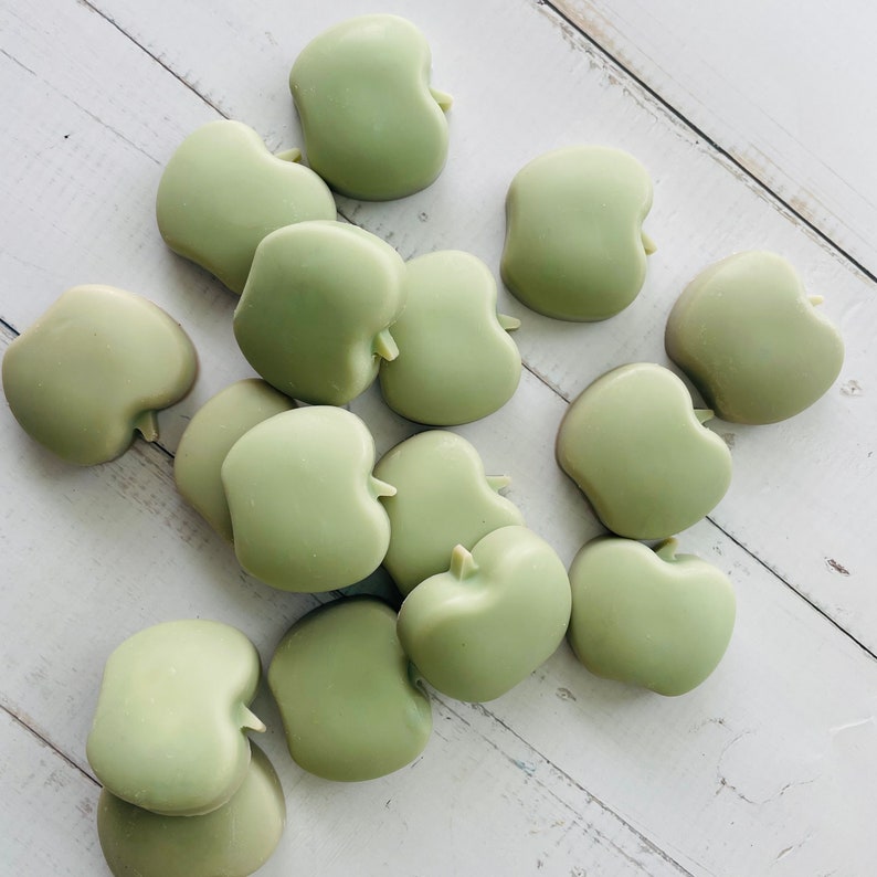 May include: A group of 15 green apple-shaped wax melts. The wax melts are arranged in a pile on a white wooden surface.