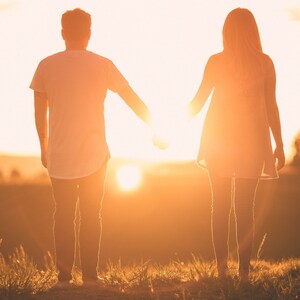 May include: Two individuals stand side by side in silhouette against a bright golden sunset. One wears a short-sleeved t-shirt and long pants, while the other wears a sleeveless dress. They are holding hands, with the sun positioned between them.