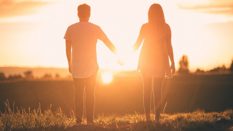 May include: Two individuals stand side by side in silhouette against a bright golden sunset. One wears a short-sleeved t-shirt and long pants, while the other wears a sleeveless dress. They are holding hands, with the sun positioned between them.