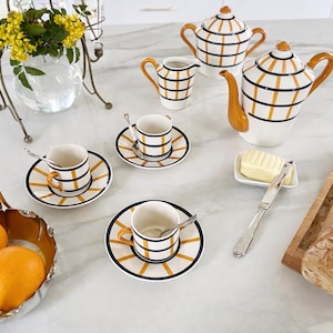 Peut inclure: Service à thé en céramique blanche avec un motif à rayures noires et jaunes, comprenant une théière, une crémière, des tasses et des soucoupes. Un panier argenté contient deux oranges. Un pain et un beurrier sont également présents.