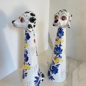 Peut inclure: Deux figurines de chiens en céramique avec des corps blancs, des taches noires sur la tête et des yeux orange. Les corps coniques allongés sont ornés de motifs floraux bleus peints à la main, d'accents jaunes et de fines lignes noires.