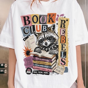 Könnte beinhalten: Weißes T-Shirt mit einer bunten Grafik, die einen Waschbären mit Brille und Nietenkragen zeigt, der einen Stapel Bücher liest. Der Text "BOOK CLUB REBELS" und "READ. THINK. REBEL." sind ebenfalls abgebildet.