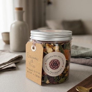 Può includere: Un barattolo in plastica trasparente riempito di pot-pourri essiccato, con un coperchio argentato e un cartellino in carta marrone legato con uno spago. Il cartellino riporta Essenza di Fico, FOGLIE DI FICO, ROSA e SPEZIE. Un'etichetta circolare mostra un fiore rosa e il testo OFFICINA DEL POT POURRI HOME FRAGRANCES MADE IN ITALY.