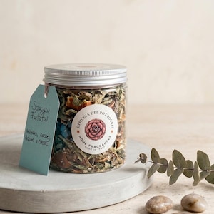 Könnte beinhalten: Ein durchsichtiges Kunststoffglas gefüllt mit getrocknetem Potpourri, verschlossen mit einem silbernen Deckel. Ein rundes Etikett zeigt eine rosa Blumenillustration und den Text Officina del Pot Pourri Home Fragrances Made in Italy. Ein hellblaues Schild trägt die Aufschrift Spiaggia Fruttata Ananas, Cocco, Agrumi e Spezie.