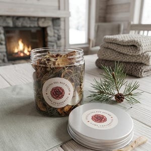 Può includere: Un barattolo di vetro trasparente pieno di pot-pourri essiccato, con coperchio argentato e cucchiaio di legno. Il barattolo ha un'etichetta con la scritta "Officina Del Pot Pourri Home Fragrances Made in Italy". Un rametto di pino e una pila di asciugamani sono sullo sfondo.