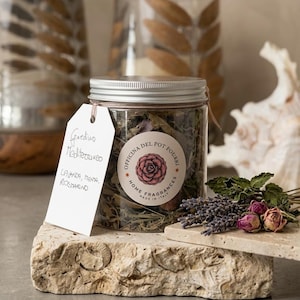 Può includere: Un barattolo in plastica trasparente riempito di pot-pourri essiccato, con un tappo a vite argentato e un'etichetta rotonda con l'illustrazione di una rosa rosa e il testo Officina del Pot Pourri Home Fragrances Made in Italy. Un cartellino bianco riporta Giardino Mediterraneo Lavanda, Menta, Rosmarino.