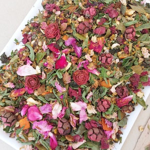 Könnte beinhalten: Eine weiße Keramik Schale gefüllt mit einer Mischung aus getrockneten Blumen, Kräutern und Fruchtschalen. Die Mischung enthält rosa Rosenblätter, rote Rosenknospen, grüne Blätter und Orangenschalen.