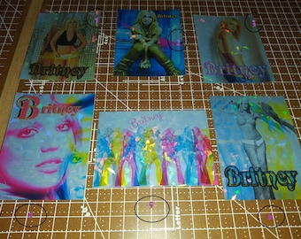 May include: Six rectangular holographic stickers featuring various photographic portraits and full-body images of Britney Spears. Each sticker displays the name Britney in different fonts and colors, with a shimmering, multi-colored prismatic finish.
