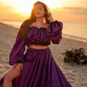 May include: A woman wearing a purple satin top and skirt with long sleeves. The top is off the shoulder and the skirt is flowing. She is standing on a beach with the sun setting in the background.